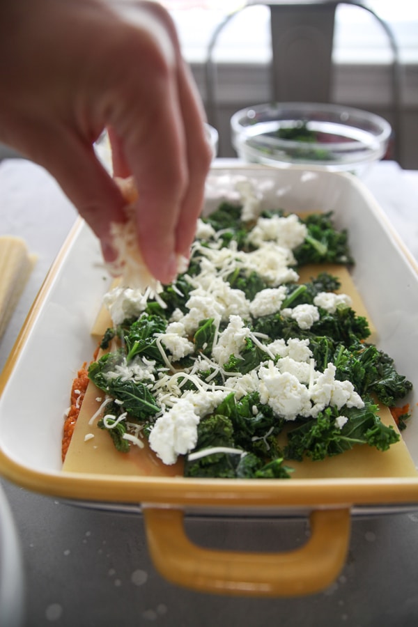 kale and goat cheese layer in a yellow casserole dish with a hand sprinkling shredded cheese