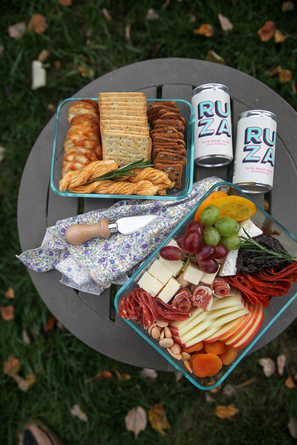 cheese and fruit in a glass container with crackers and cans of wine