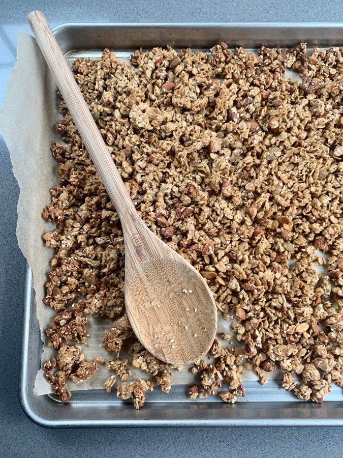 Overhead shot of Crunchy Tahini Granola on stainless steel sheet pan with parchment paper and wooden spoon.