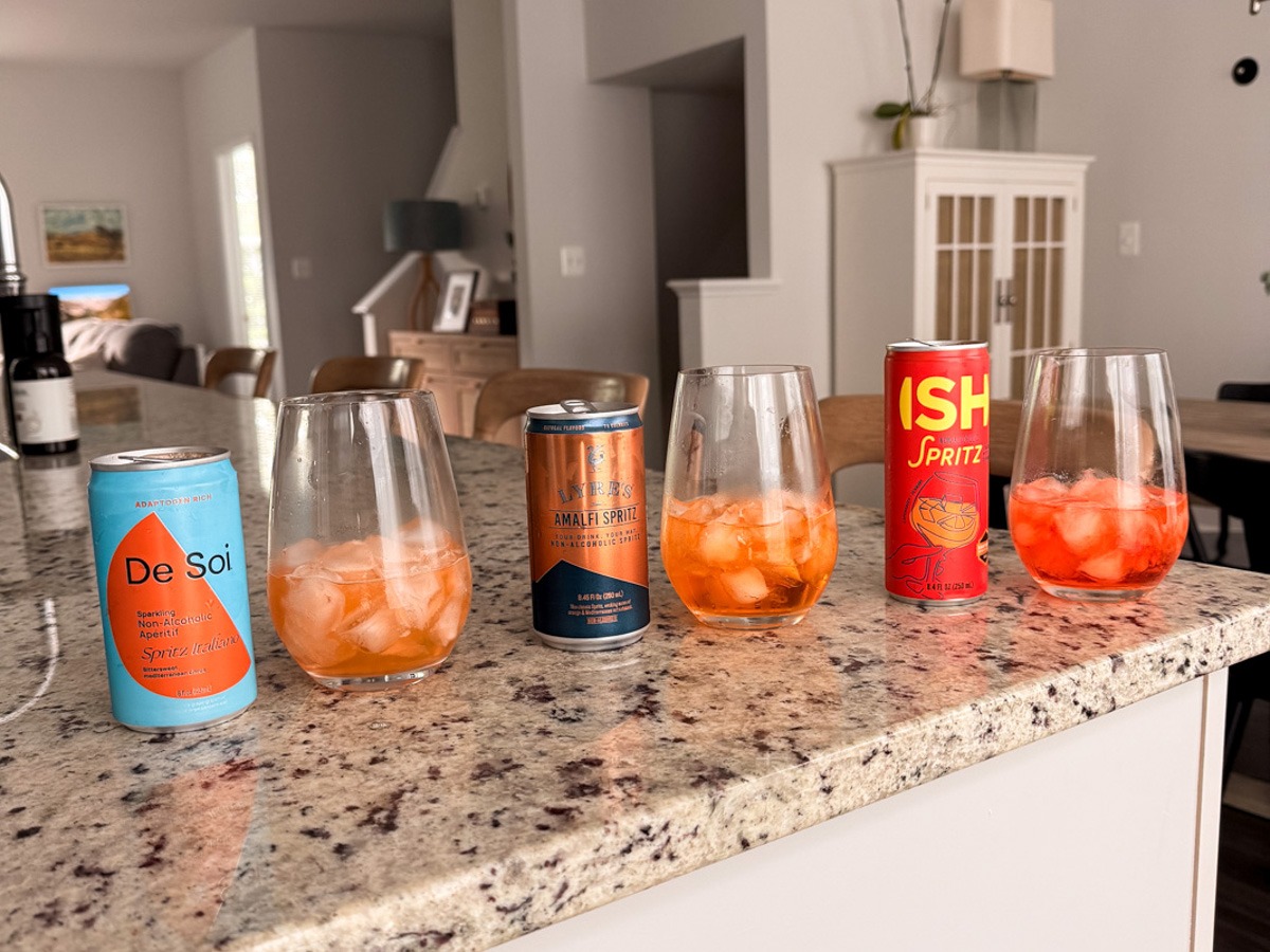 a row of nonalcoholic aperol spritzes: de sio, Lyre's, Ish all poured in wine glasses with ice in Kath's kitchen